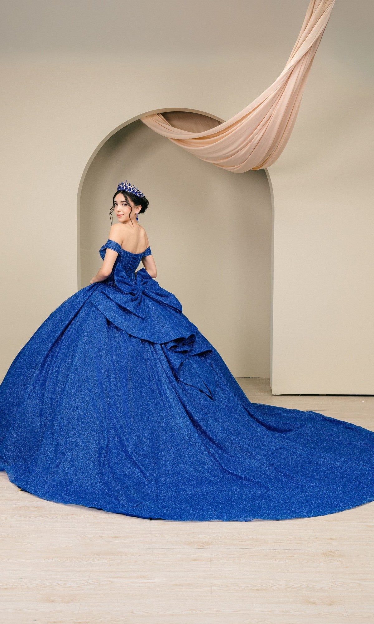 A woman in the Quinceanera Dress 8460JQ by Cinderella Couture—an off-the-shoulder blue glitter gown with a long train and matching crown—stands before a neutral arched backdrop with draped fabric overhead.